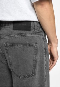 Jean en denim gris avec une étiquette en cuir embossée du mot « EIGHTYFIVE » à l'arrière de la taille, avec des coutures visibles et des poches arrière.