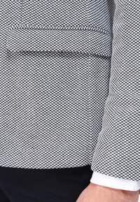 Näruppfattning av en persons överkropp som bär en strukturerad kavaj med svart och vitt mönster, med en fickklaff och en vit skjortärm synlig.