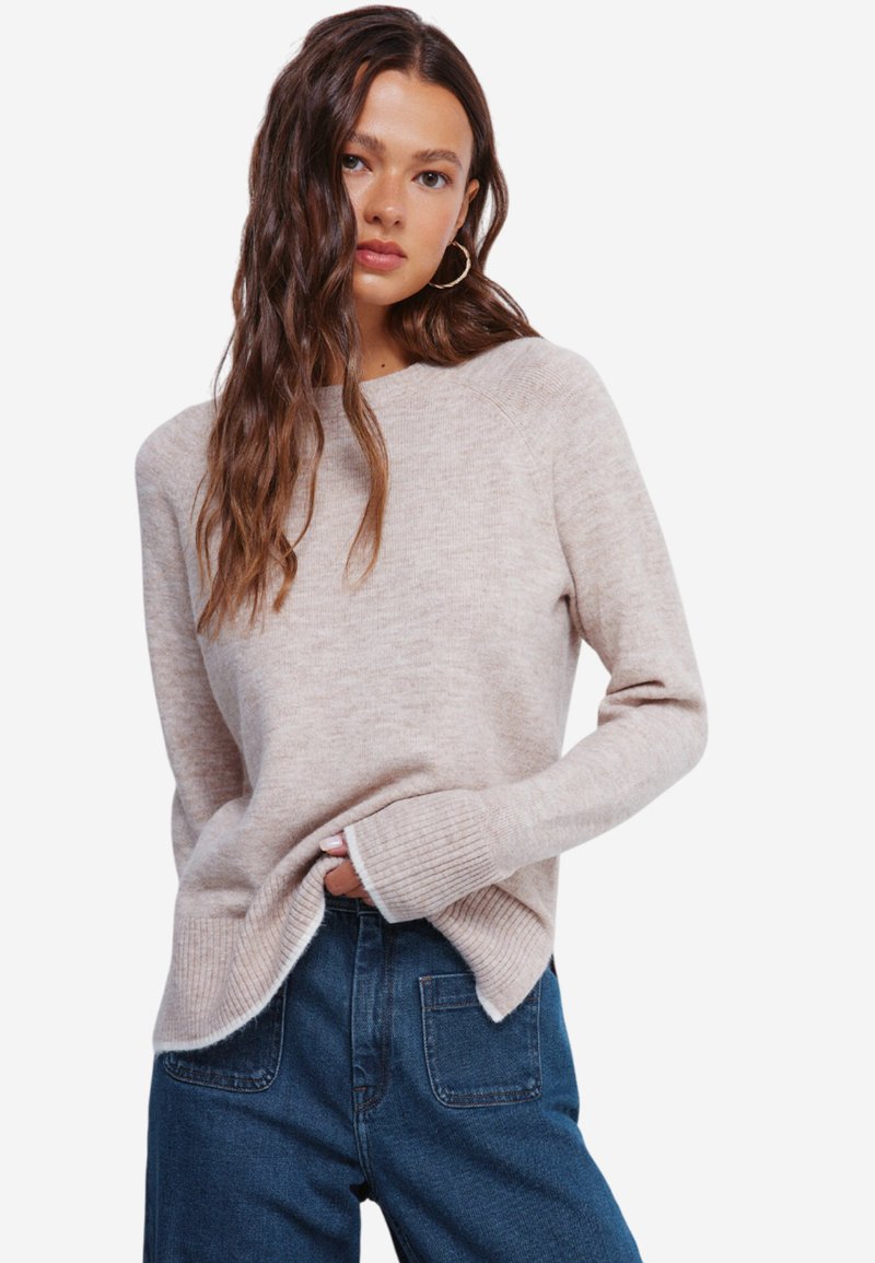 Maglione a maglia color beige chiaro con collo girocollo, polsini a coste e spacchi laterali, abbinato a jeans a vita alta di colore blu scuro. Dettaglio del tessuto testurizzato visibile.