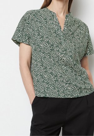Femme portant un chemisier vert manches courtes avec un motif abstrait blanc et un pantalon noir, debout les mains dans les poches.
