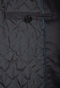 Svart kviltad tyg med ett diamantmönster, med teal-färgad söm och en svart knappdetalj på vänster sida. Slät textur.