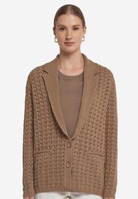 Brun stickad cardigan med ett strukturerat mönster, en v-formad krage, två framfickor och två knappstängningar. Bärs över en matchande topp.