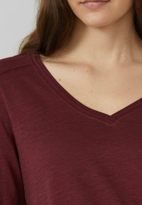 Burgundia V-kaelusega topp, mis on valmistatud pehmest, tekstuuriga kangast, omades lõdvalt istuvat viisi lühikeste varrukatega, millel on õrnad õmblusdetailid.