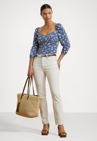 Bluse mit blauen Blumenmuster und Puffärmeln, kombiniert mit cremefarbenen Hosen. Accessoires bestehen aus einer geflochtenen Strohtasche und braunen Plateauschuhen.