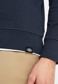 Laivastonsininen collegepaita ribbineuloksella ja resoreilla, jonka helmassa on musta Dickies-logo. Paita on yllään vaaleiden housujen päällä.