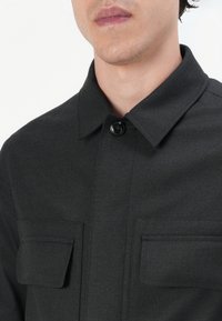 Black button-up shirt with a collar, featuring two chest pockets and a textured fabric. Close-up view, showcasing the material and button detail.