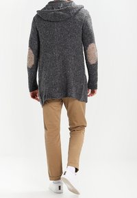 Šedý pletený cardigan s kapucí a záplatami na loktech, nošený přes béžové kalhoty, představující texturovanou tkaninu a volný střih.