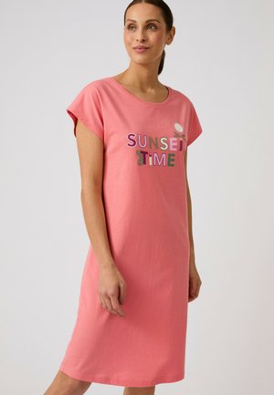 Mujer vestida con un vestido rosa coral de manga corta y "SUNSET TIME" texto colorido en la parte delantera, de pie frente a un fondo claro y liso.