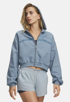Vrouw draagt een blauwe cropped hoodie van Under Armour met capuchon omhoog en bijpassende lichtblauwe sportshorts.