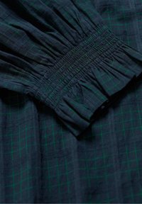 Tmavomodrá tkanina so zelenými mriežkovými líniami, ktorá má detail s gumičkou na manžete rukáva.