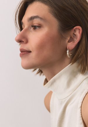 Young woman in white sleeveless turtleneck wearing gold hoop earrings with round details, looking to the left against a plain background.