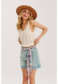 Svetlo krémová pletená top bez rukávov s výstrihom do V, skombinovaná so svetlomodrými denimovými šortkami s kvetinovým potlačou a viazaním opasku. Hnedý široký klobúk.