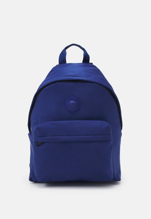 Plavi platneni ruksak zaobljenog oblika, s prednjim džepom i ručkom na vrhu. Ima kružni logo i crne detalje na zatvaraču.