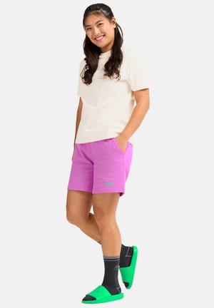 Losse pasvorm, crèmekleurig T-shirt; roze korte broek met zakken; groen accentlogo; grijze sokken; groene slidesandalen. Zachte katoenmix materialen.