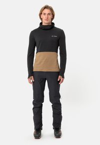 Haut à manches longues noir et beige, avec un col haut, un tissu texturé et une coupe ajustée. Associé à un pantalon sombre et des chaussures de ski.