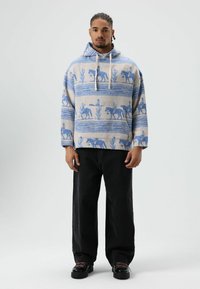 Wegwerpsweater in lichtgrijs met blauwe, met paarden en cactussen bedrukt patroon. Losse pasvorm, met trekkoord en lange mouwen. Gecombineerd met zwarte broek.