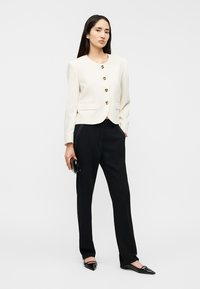 Women's cream blazer with gold buttons, paired with black trousers and pointed flats, featuring a structured design and smooth texture.