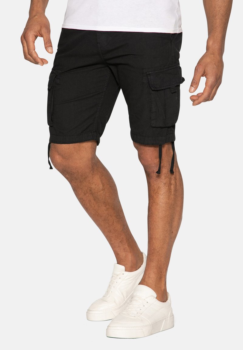 Schwarze Cargo-Shorts mit seitlichen Taschen, verstellbaren Kordeln am Saum und einem geraden Schnitt. Kombiniert mit weißen Sneakers.