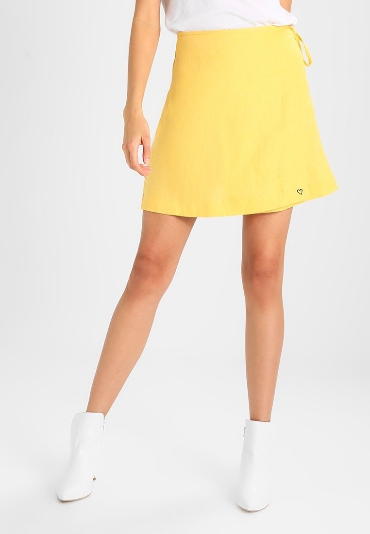 Yellow linen A-line mini skirt with a tie waist, featuring a small heart embroidery detail near the hem. Smooth texture and lightweight design.