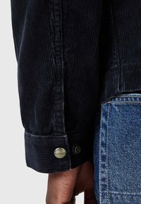 Cămașă din cordul negru, cu o textură în dungi, mansete cu nasturi și un accent vizibil cu logo. Asortată cu țesătură din denim de culoare albastru deschis.