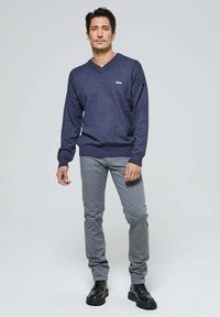 Pull en maille navy avec col en V, associé à un pantalon slim-fit gris clair. Des bottes noires à lacets complètent la tenue. Accent simple de logo sur le pull.