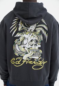 Felpa nera con cappuccio, con una grafica sul retro che raffigura un teschio, un serpente e un'aquila in colori vivaci, accompagnata dal testo "Ed Hardy".