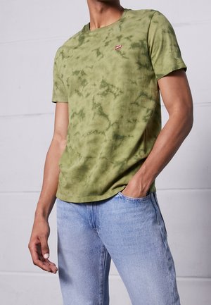 Homme portant un T-shirt à manches courtes tie-dye vert avec un petit logo Levi's rouge sur la poitrine, et un jean bleu clair, la main dans la poche.
