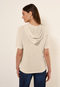 Hoodie corto a maniche corte beige chiaro realizzato in tessuto testurizzato, con cappuccio drappeggiato e polsini a costine, abbinato a jeans in denim scuro.