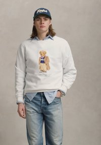 Giovane uomo con berretto blu e felpa bianca con una grafica di orsetto, camicia azzurra chiara sotto e jeans azzurri chiari, in piedi contro uno sfondo grigio.