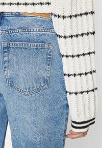 Vitt stickat tröja med svart rutmönster, kombinerat med högmidjade ljusblå denimjeans som har en enda bakficka och synlig sömnad.