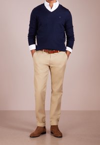Man wearing navy V-neck sweater over white shirt, beige pants with brown belt, and brown suede boots, standing with hands in pockets.