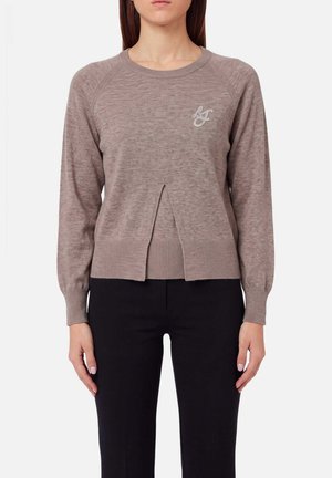 Donna che indossa un maglione in maglia color taupe con maniche lunghe, spacco frontale e logo ricamato, abbinato a pantaloni neri su sfondo bianco.