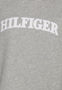 Světle šedá tkanina s bílým vyšívaným textem "HILFIGER" v obloukovém, vysokoškolském stylu.