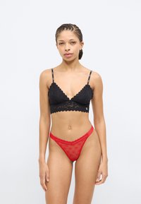 Bralette en dentelle noire à bords festonnés, avec de fines bretelles. Associé à un string rouge transparent, texturé avec une ceinture en dentelle.