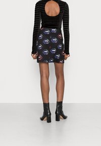 Kvinna i en svart långärmad topp med öppen rygg, en kort svart kjol med färgglada läpptryck och svarta högklackade ankelskor.