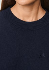 Granatowy sweter z dzianiny z prążkowanym, okrągłym dekoltem. Posiada subtelną fakturę i małe logo na lewej stronie klatki piersiowej.