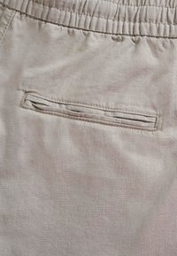 Tissu beige avec une ceinture élastique et une poche horizontale style fente cousue dans le matériau.