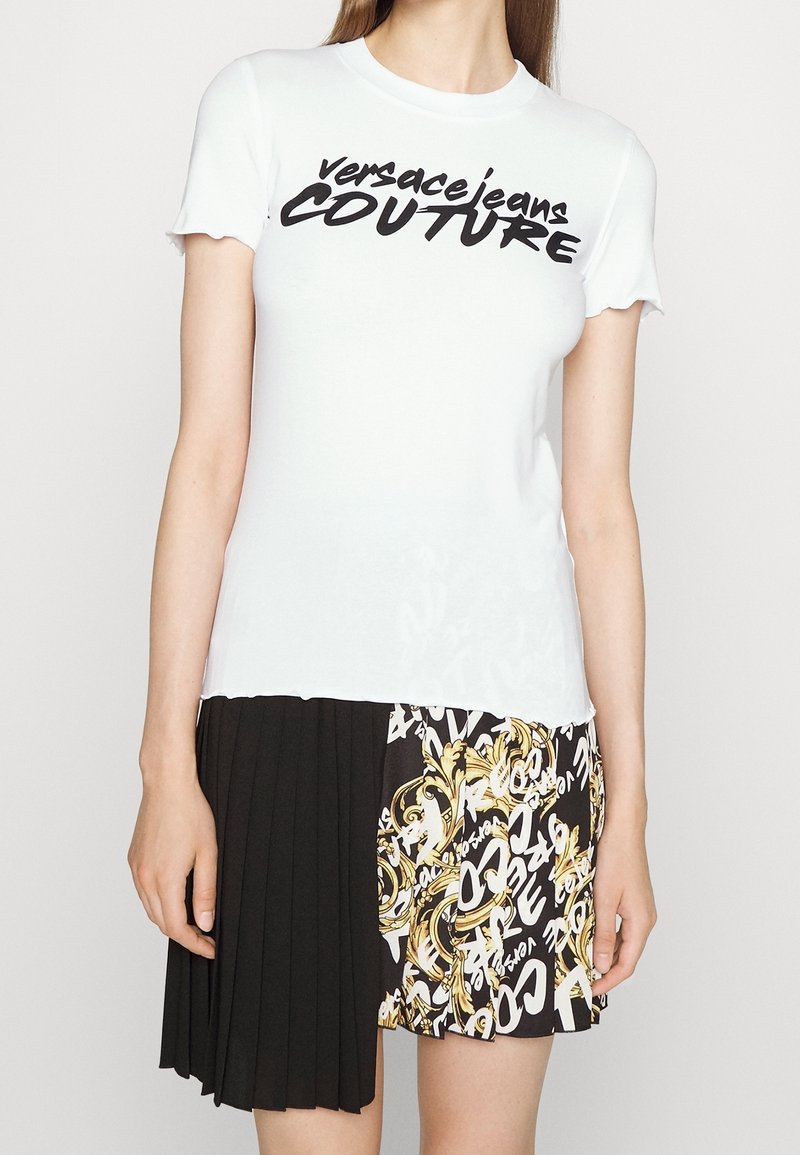 Vit bomulls t-shirt med korta, rynkiga ärmar och svart text "versace jeans COUTURE"; kombinerat med en plisserad svart och mönstrad kjol.