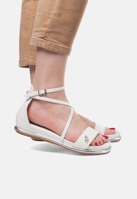 Bílé kožené sandály s křížovými kotníkovými pásky, otevřenou špičkou, stříbrnými akcenty a plochou podrážkou. Logo umístěné na pásku.