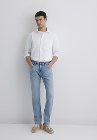 White button-up shirt, light blue jeans, and beige sneakers. Model stands with hands in pockets, against a plain white background.