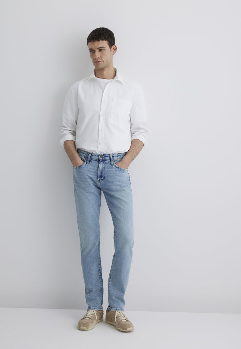 White button-up shirt, light blue jeans, and beige sneakers. Model stands with hands in pockets, against a plain white background.