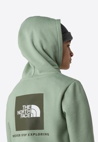 Lys grønn hettejakke med en stor firkantet logo i mørkegrønt og hvitt på baksiden, med myk tekstur og hette med snøring.