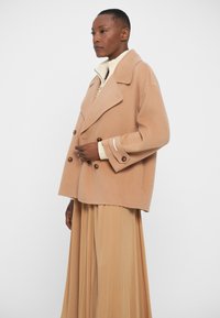 Beige double-breasted wool coat with large buttons, peaked lapel, and wide sleeves, paired with a light cream knitted collar and pleated skirt.