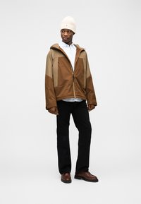 Καφέ και καμηλό μπουφάν με κουκούλα και φερμουάρ μπροστά, φορεμένο πάνω από ανοιχτό μπλε πουκάμισο με κουμπιά, μαύρο παντελόνι και καφέ παπούτσια. Μπεζ σκούφος.