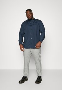 Navy button-up shirt with a stand collar, long sleeves. Paired with light gray plaid trousers and black lace-up shoes. Flat texture.