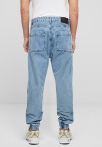 Pantaloni jogger din denim de un albastru deschis, cu manșete elastice și două buzunare pe spate. Prezintă un eticheta neagră la talie, un design cu picioare drepte și o textură netedă.
