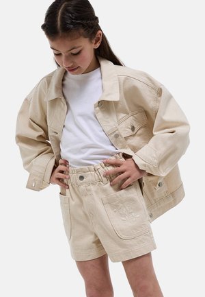 Fille portant une veste en jean beige et un short avec broderie florale, regardant vers le bas, les mains sur les hanches, sur fond blanc.