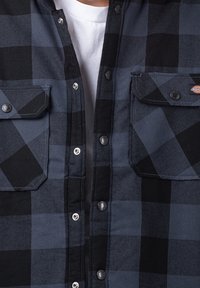 Svart och mörkblå rutigt flanellskjorta med två bröstfickor, tryckknappar och en bomullstextur. Har kontrasterande svarta detaljer.