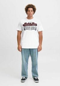 Bijela majica s crvenim i crnim natpisom "Jack & Jones Superior Quality Denim Goods." Uparena sa svijetloplavim trapericama i crnim tenama.