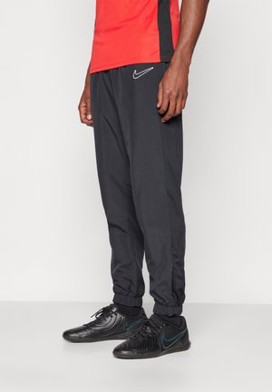Homme portant un pantalon de survêtement Nike noir, des baskets Nike noires avec un swoosh bleu sarcelle, et un maillot de sport rouge sur un fond blanc uni.